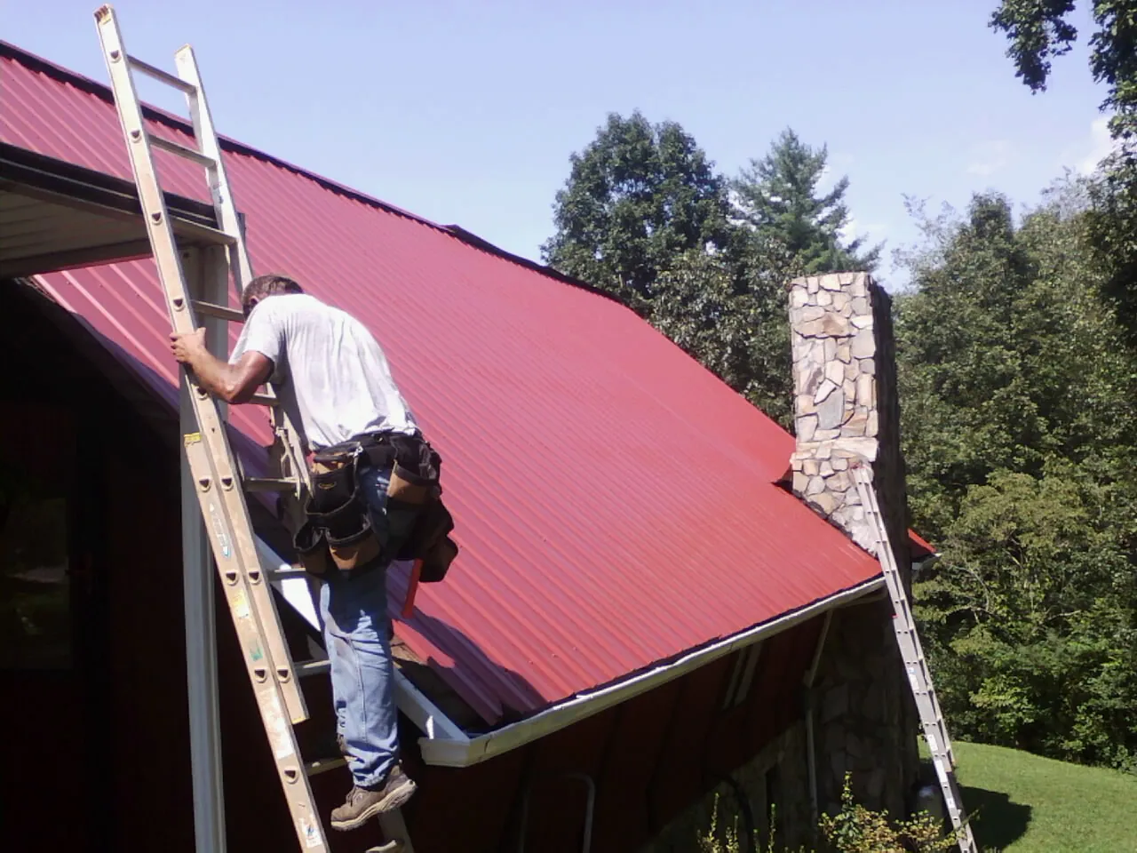 Roofer on a ladder beside a red metal roof and stone chimney — Soffit & Fascia Repair by Roofing Brothers in Guysville